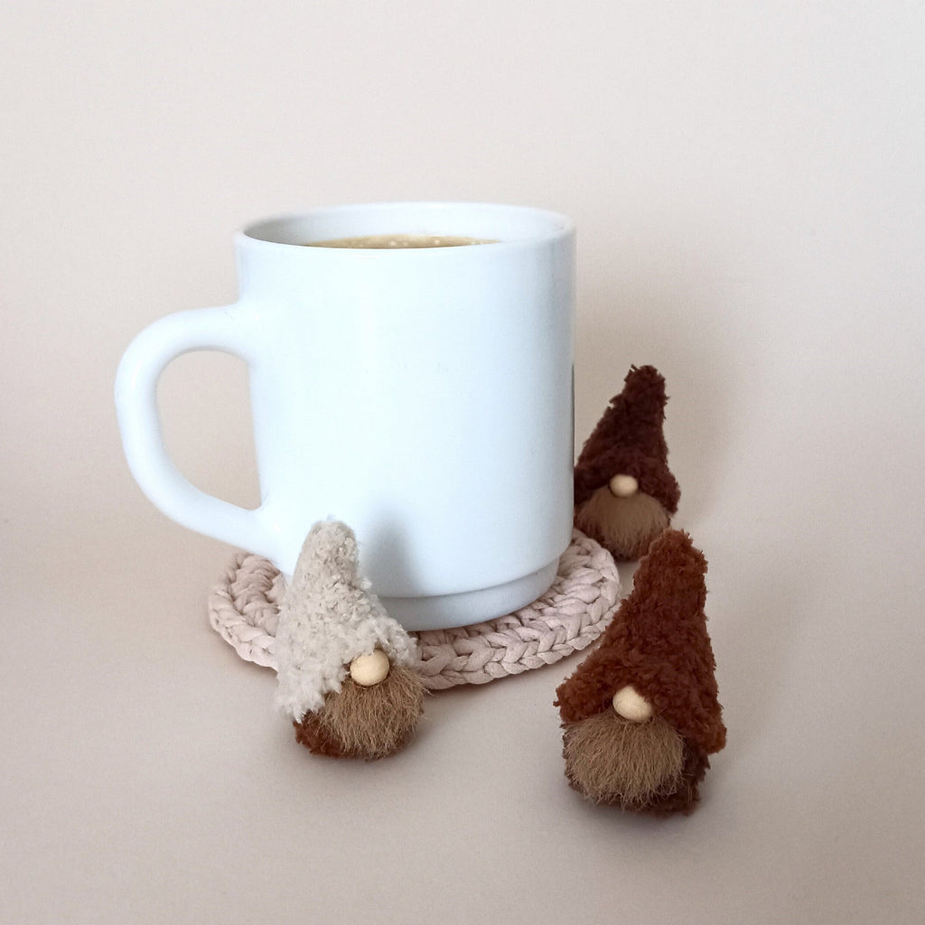Three tiny handmade coffee gnome plushies in cream, cappuccino, and espresso brown standing around a white coffee mug on a neutral background. Cozy office desk accessories and cute coffee station decor for coffee lovers.