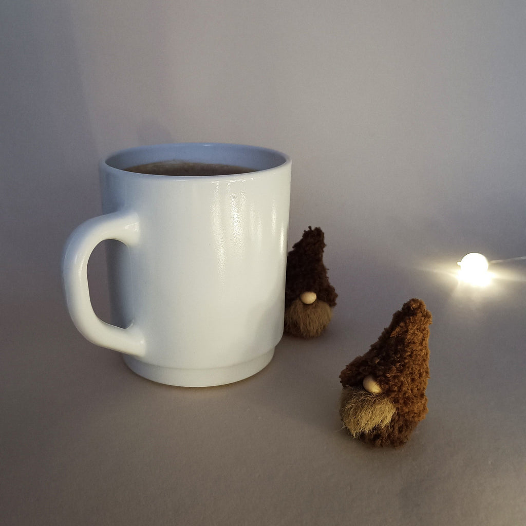 Two small handmade coffee gnome plushies next to a white mug, illuminated by a warm fairy light in a cozy setting. Aesthetic winter holiday desk decor and unique minimalist Christmas stocking stuffer ideas.
