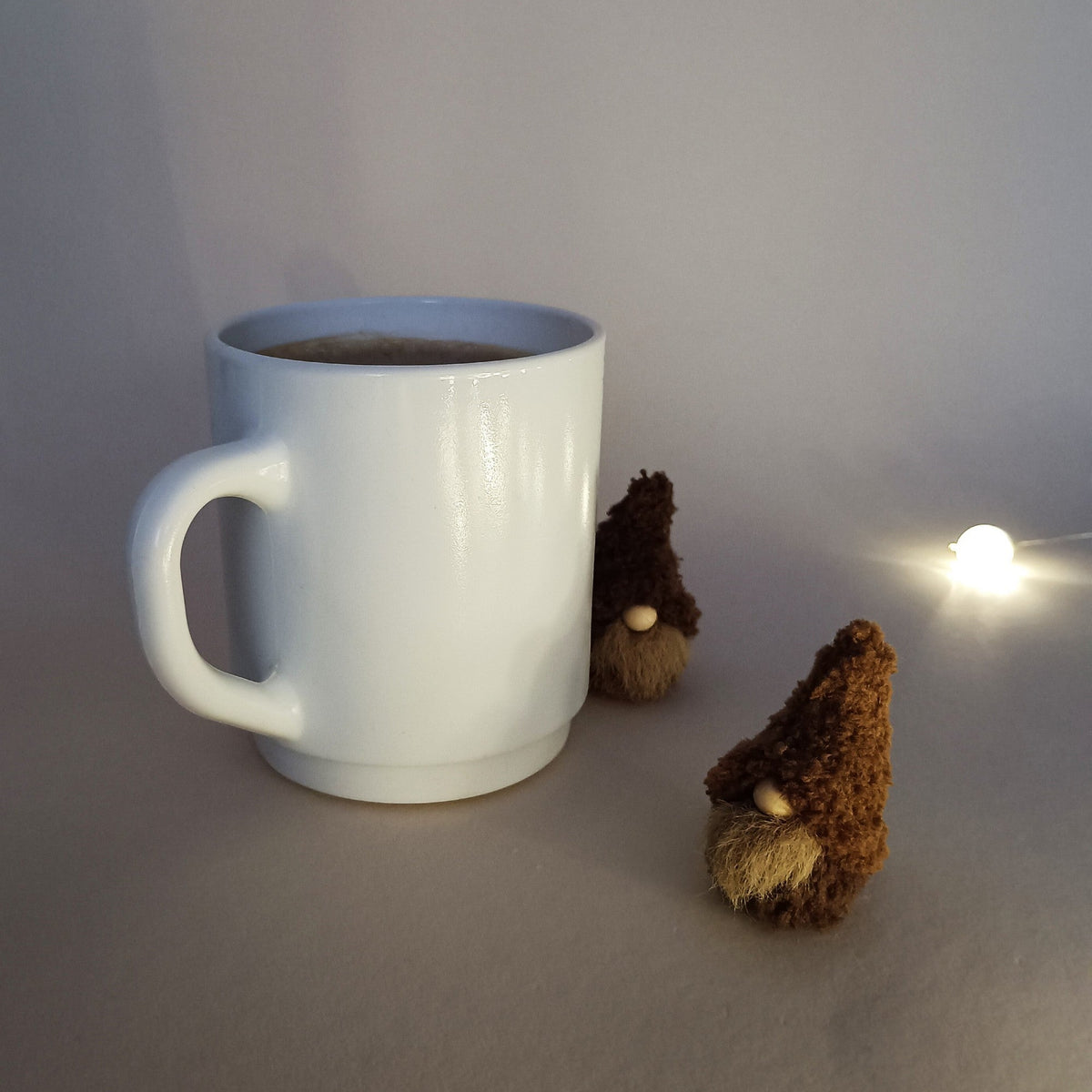 Two small handmade coffee gnome plushies next to a white mug, illuminated by a warm fairy light in a cozy setting. Aesthetic winter holiday desk decor and unique minimalist Christmas stocking stuffer ideas.