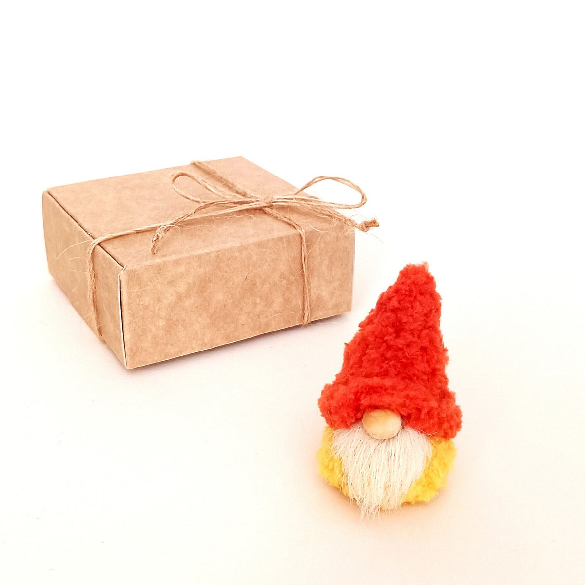 A tiny handmade pumpkin spice gnome plush standing next to a kraft paper gift box tied with a twine bow. Aesthetic minimalist packaging for a small autumn coworker gift and unique thinking of you treat.