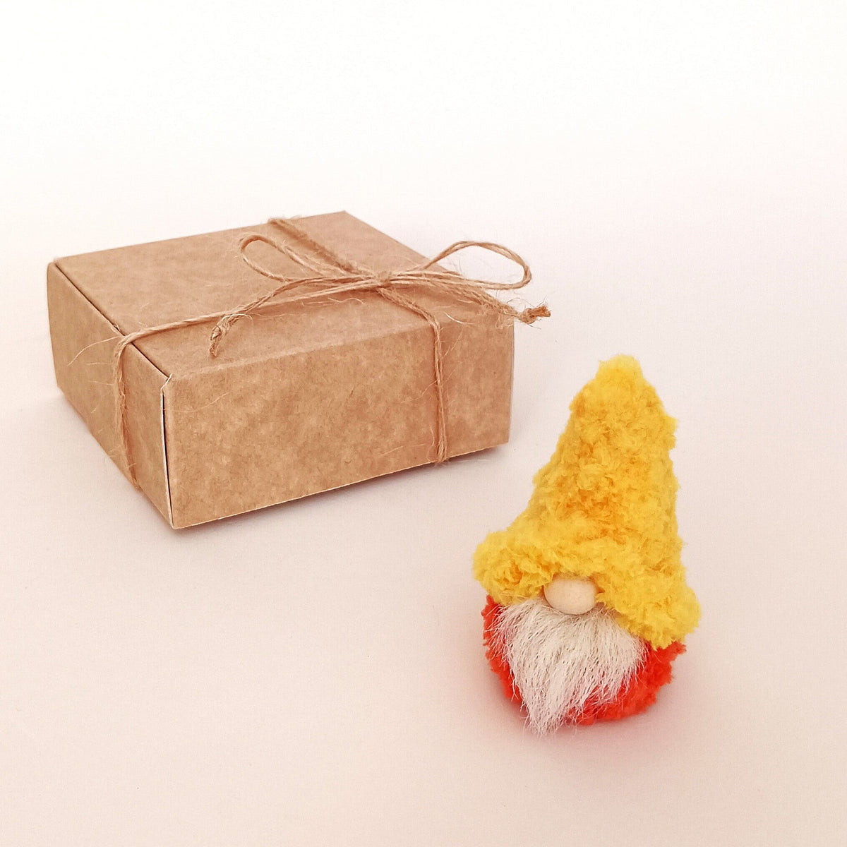 Golden honey yellow gnome style option standing next to a kraft paper gift box tied with a twine bow. Miniature primitive autumn desk pet variation and unique small holiday gift box idea for coworkers.