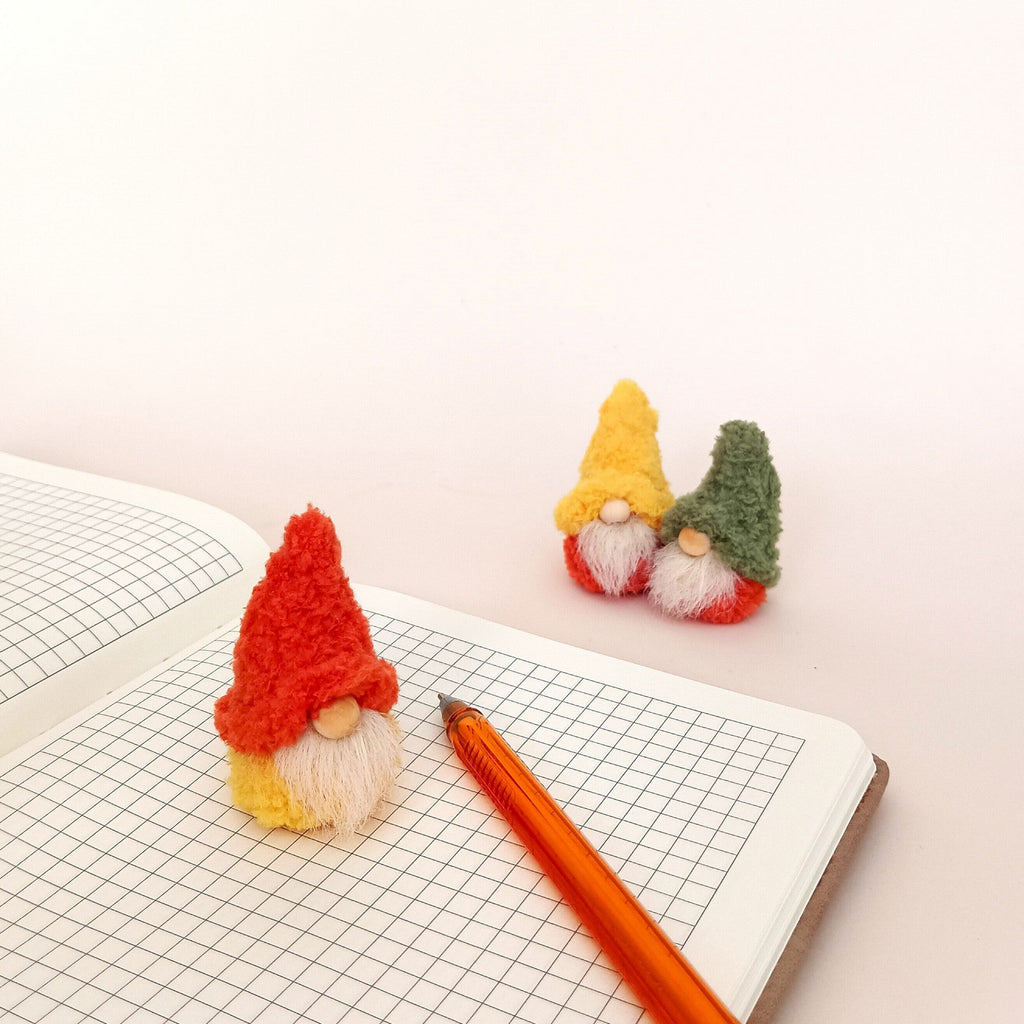 A miniature fall desk decor set featuring one pumpkin spice gnome on an open paper notebook next to an orange pen, with two tiny gnomes sitting in the background. Cozy minimalist workspace accessories and aesthetic home office gifts for adults.
