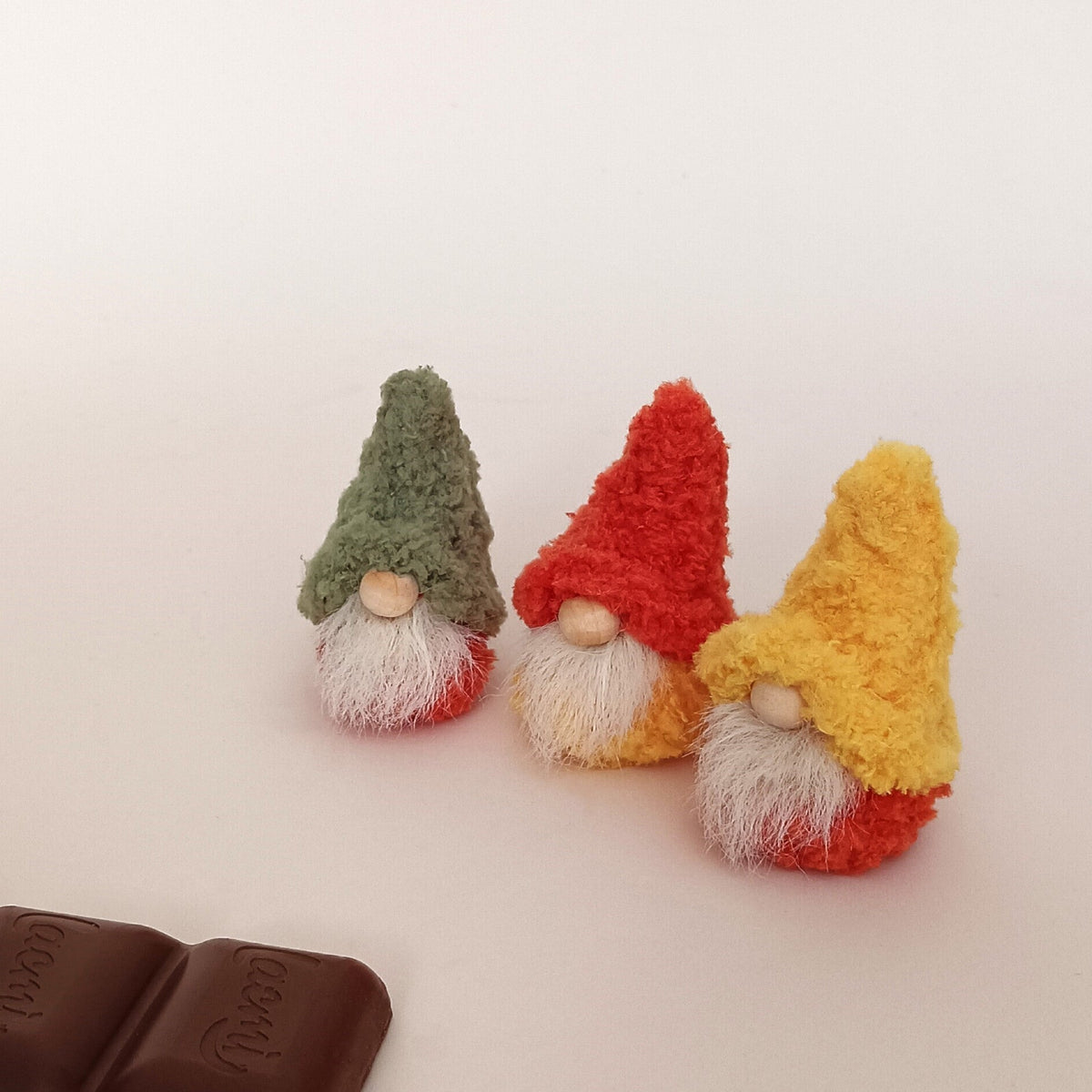 Three miniature handmade autumn gnomes in green, orange, and yellow boucle hats sitting together next to a dark chocolate bar. Cute hygge desk pet variations and cozy fall office workspace companion gifts for adults.