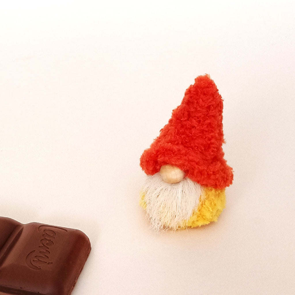 A single miniature handmade pumpkin spice gnome plush sitting on a clean light background next to a dark chocolate bar. Cute hygge office desk pet and a cozy small autumn themed thank you gift idea for coworkers.
