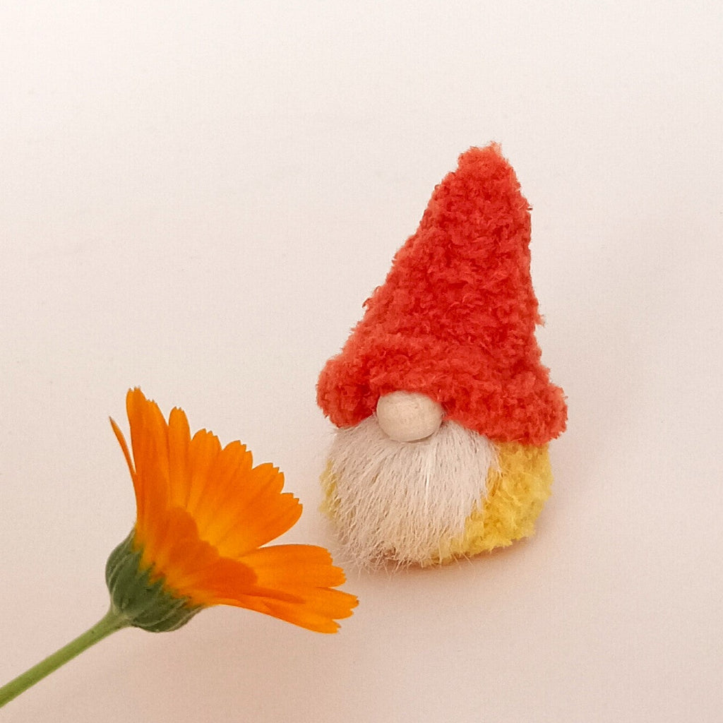 A miniature handmade orange pumpkin spice gnome standing next to an orange calendula flower on a clean white background. Cute primitive desk pet ornament and unique small autumn themed thank you gift idea for coworker or coffee lover.