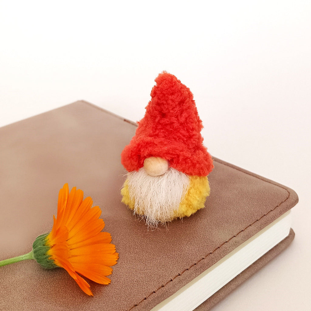 A tiny handmade pumpkin spice gnome plush sitting on a brown leather planner next to a vibrant orange calendula flower. Cozy fall desk aesthetic decor and minimalist autumn office workspace companion for adults.