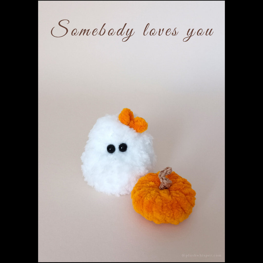 A 5x7 printable minimalist greeting card featuring a tiny white ghost handmade from fluffy plush crochet yarn with a vibrant orange bow and a small textile pumpkin. Solid light beige studio background with elegant empty space and clean chocolate brown typography that reads Somebody loves you at the top in a luxury wabi-sabi style.